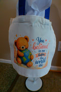 White canvas tote bag with blue handles and teddy bear hugging the world design