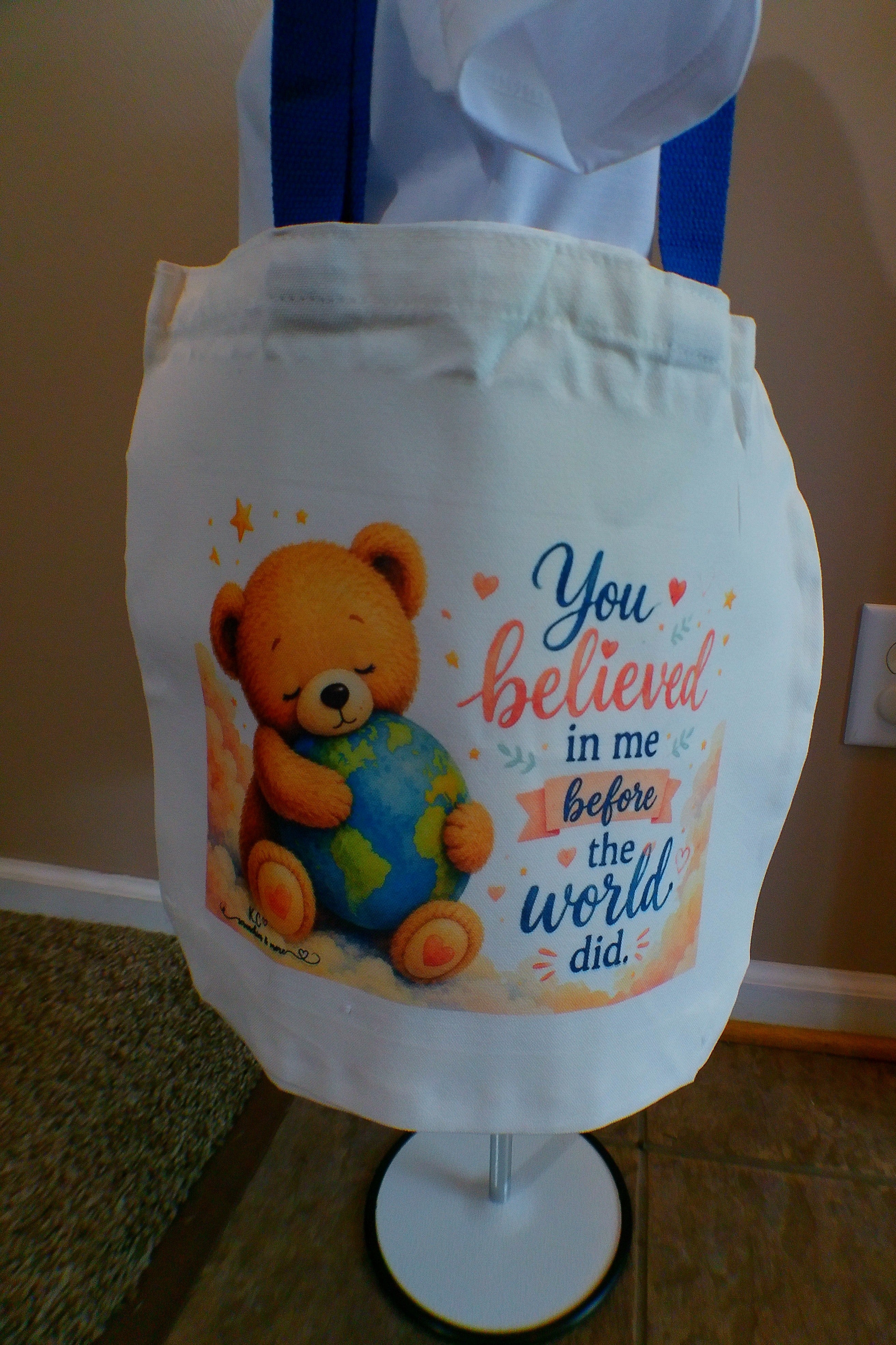 White canvas tote bag with blue handles and teddy bear hugging the world design